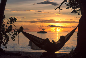 Beach vacations, silhouette of a woman reading in hammock at sunset on tropical island.