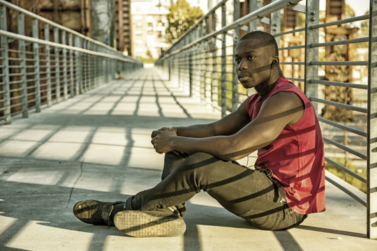 Portrait of a sexy young black man in urban environment wearing red sleeveless shirt, sitting against railing