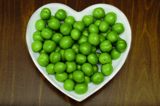 Heart form white bowl  full of Fresh green Plums Greengages on wooden Table. Georgian Fruit Alucha