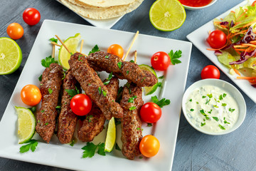 Homemade Kofta kebabs on skewers with pita, lime, vegetables, sweet chili and yogurt sauce on white plate.