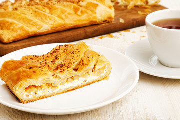 Piece of homemade puff braided pie from with cottage cheese, apples and raisins on white wooden table.