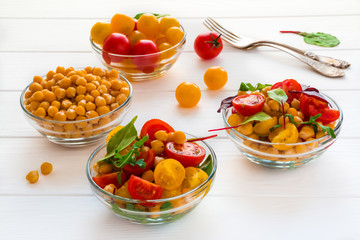 Chickpea salad with fresh red and yellow cherry tomatoes and mix of green salad leaves in glass bowls on the white wooden table.