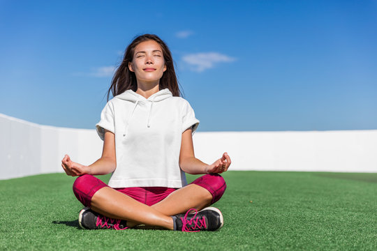 Yoga fitness woman doing summer meditation outdoor. Happy asian athlete meditating before strength training workout sitting in lotus position on outside grass gym floor. Active living.