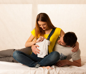 Beautiful coule  - young man and pregnant woman resting on bed holding babys dress