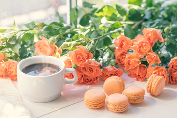 Orange mango or citrous macaroons, cup of coffee and fresh little roses on light wooden background. Sunlight. Coloring and processing photo with light vintage style. Shallow depth of field.