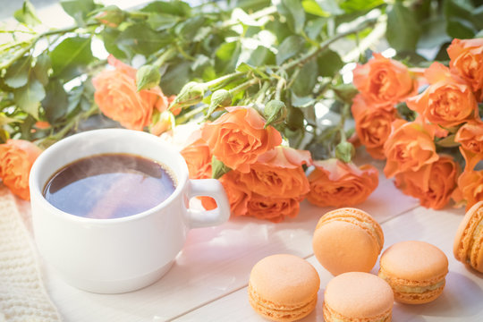 Orange mango or citrous macaroons, cup of coffee and fresh little roses on light wooden background. Sunlight. Coloring and processing photo with light vintage style. Shallow depth of field.