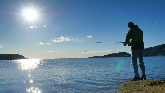The active man is fishing on sea from the rocky coast. Fisherman check pushing bait on the fishing line, prepare rod and than throw lure into peacefull water. Fisherman silhouette at sunset