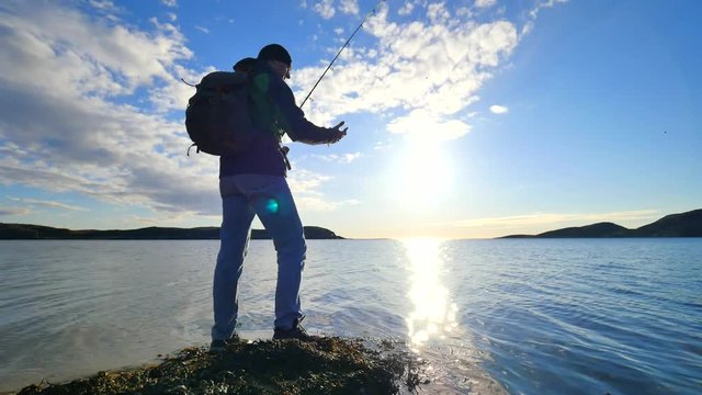 The fisherman with green sporty backpack is checking fishing equipment.  Man prepare pushing bait on fishing line and throw lure far into calm sea water. Fisherman enjoy afternoon sport activities.