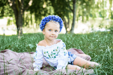 Cute smiling little girl with blue eyes in embroidered shirt with blue wreath of wild flowers on the head sitting in the grass on a sunny summer day 
