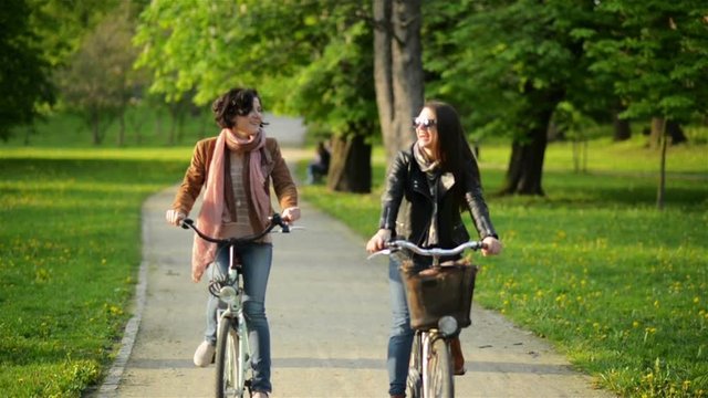 Two Active Girl are Riding Together in the City Park. Smiling Young Women with Bicycles Outdoors.