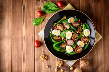 Fresh vegetable salad with spinach, cherry tomatoes, quail eggs, pomegranate seeds and walnuts in black plate on wooden table.