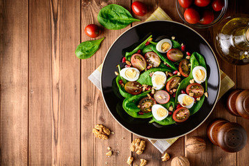 Fresh vegetable salad with spinach, cherry tomatoes, quail eggs, pomegranate seeds and walnuts in black plate on wooden table.