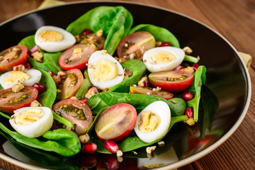 Fresh vegetable salad with spinach, cherry tomatoes, quail eggs, pomegranate seeds and walnuts in black plate on wooden table.