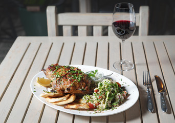 Leg of lamb baked with sauce in the white plate closeup with a side dish of rice. Rustic wooden table and a glass of red wine.