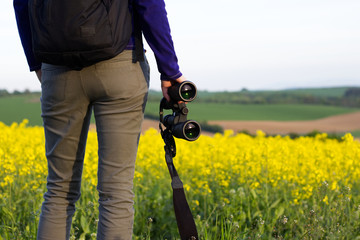 Young hiking woman watching landscape with binoculars. Tourist girl in rape field at countryside. Binoculars in a female hand.