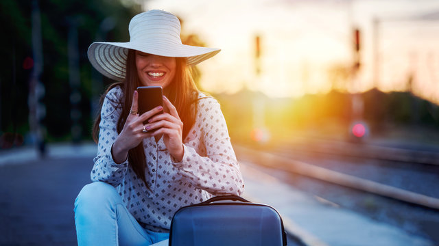 Young smiling woman with baggage on train station using smart phone