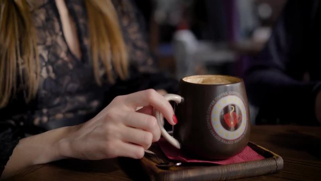 Close up of female elegant hands holding big brown ceramic mug and green decor plant in stylish glass. Woman in black lace sexy decollette dress sitting at the table with cup of coffee and speaking.