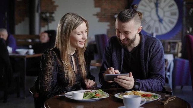 Happy couple having lunch together and looking at smartphone. Man and woman using digital gadget laughing at pictures inside. Friends in restaurant watching on display of cellphone and smiling.