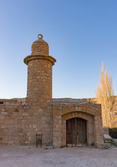 Mosque in Dana Jordan made from stones with minaret topped with crescent moon and wooden arched doorway. Background has deep blue sky and sunlit tree. 