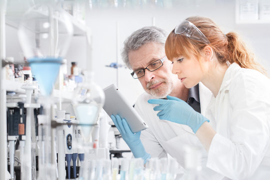 Health Care Researchers Working In Life Scientific Laboratory. Young Female Research Scientist And Senior Male Supervisor Looking Focused At Tablet Computer Screen Evaluating And Analyzing Study Data.