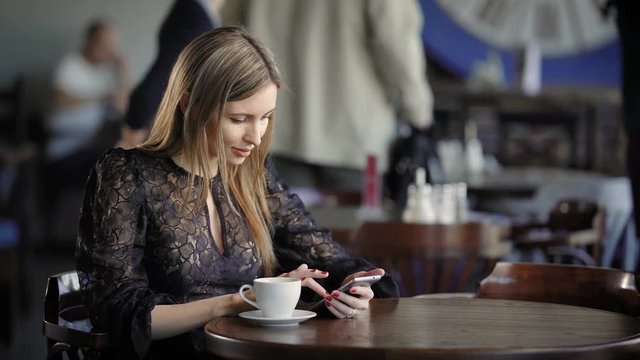 Close up of beautiful woman spending leisure time using modern smartphone and browsing Internet. Adorable lady relaxing in coffee shop during the lunch with a cup of beverage and digital device.