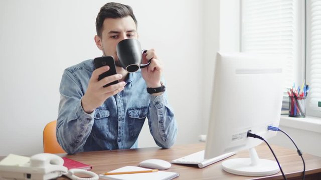 Young office worker using his phone at the office sitting at the table with computer, phone and drinking coffee or tea from his cup. Shot in 4k