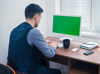 Young man office worker sitting at computer with Chromakey, looking on monitor and typing. Office concept with working environment. Back view. Copyspace on screen