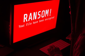 Computer screen with ransomware attacks alert in red and a man keying on keyboard in a dark room,...