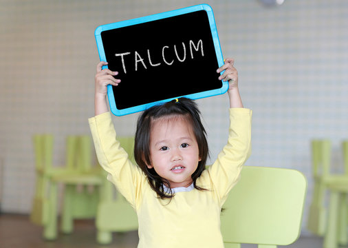Cute little child girl holding blackboard showing text " TALCUM " in kids room. Education concept.