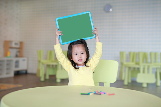 Cute asian child girl holding empty green blackboard in kids room.