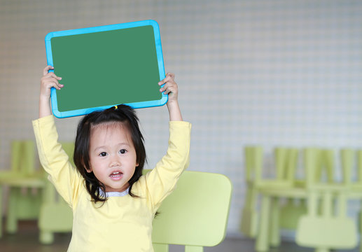 Cute asian child girl holding empty green blackboard in playroom.