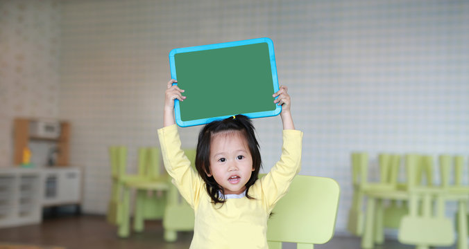 Cute asian child girl holding empty green blackboard in playroom.