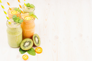 Freshly blended orange kumquat and green kiwi fruit smoothie in glass jars with straw, mint leaf, cut ripe berry, copy space. White wooden board background.