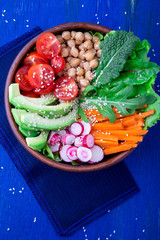 Vegan buddha bowl on blue wooden background. Bowl with carrot, lettuce, tomatoes cherry, radish, avocado and chickpea. Vegetarian, healthy, detox food concept. Top view.