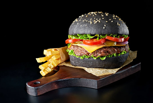 Black burger with french fries on wooden cutting board isolated on black background.