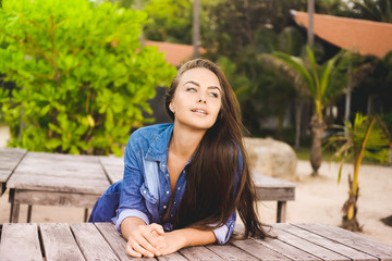 close-up portrait of a beautiful young brunette girl with long hair on a background of green tropical summer, lifestyle, posing and smiling, on a wooden platform in a denim jacket