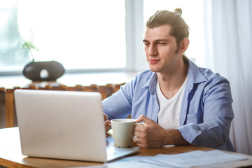 Young man holding mug, sitting at the table and looking to the laptop screen