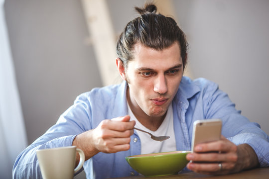 Young man eating corn flakes with milk and and looking to thesmartphone screen