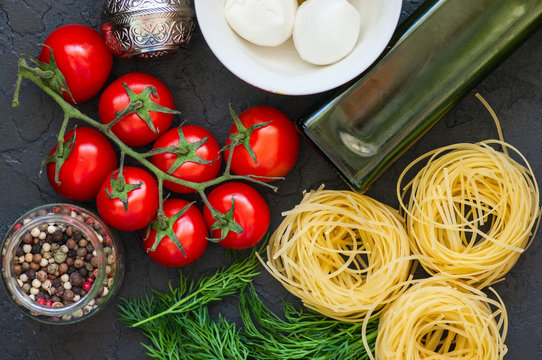 Ingredients for italian dinner. Olive oil, dill, cherry tomatoes, spices, mozzarella and italian whole grain pasta on a black slate background. Top view and copy space.