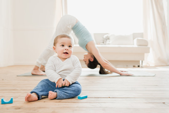 Young Woman In Downward Dog Position While Her Baby Playing At Home