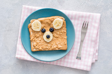 Teddy bear toast for kids. Crunchy peanut butter, banana and blueberry toast with cute teddy bear face on a blue plate over pink checkered textile. Top view. Concept of kids breakfast, food for kids
