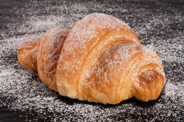 one croissant sprinkled with powdered sugar on black stone background closeup