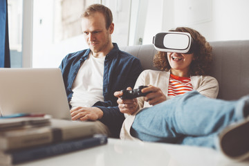 Naklejka premium Pretty couple using electronic devices. Woman playing breathtaking computer game with VR headset. Man typing on laptop. Happy family life