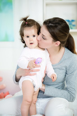 Young mom kissing her baby girl on the bed.Baby standing and looking at camera with open mouth .Shallow doff