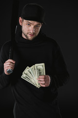young robber in zoodie with baseball bat and money isolated on black