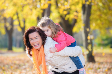 Fototapeta premium Mother giving piggy back to her little daughter in the autumn park