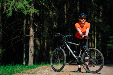 Naklejka premium Young pretty woman in helmet and sunglasses standing with bicycle on the forest road. Healthy lifestyle concept. Girl with bike.