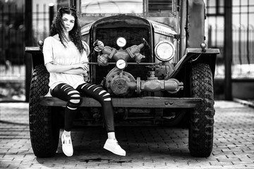 Fototapeta na wymiar Vintage firefighting truck and pretty teenager. Outdoor portrait of a beautiful young white girl with red curly hair and in black ragged jeans posing near old fire engine. Black and white.