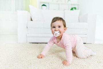 Happy baby little girl crawling with a pacifier in a mouth on the living room floor, looking at camera.Copy space