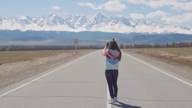 Alone woman tourist is taking pictures on smart phone while tay on the road highway with mountains on background